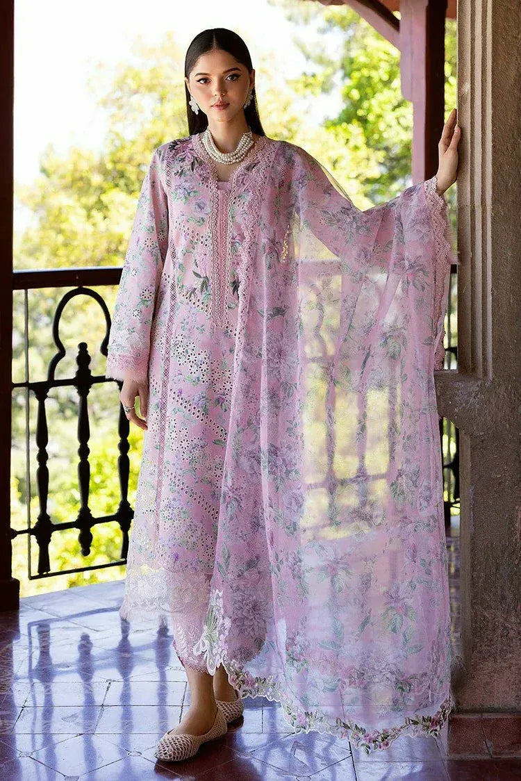 A woman models a light pink embroidered traditional outfit with a matching sheer dupatta on a sunlit balcony. The outfit features intricate floral embroidery and lace detailing.