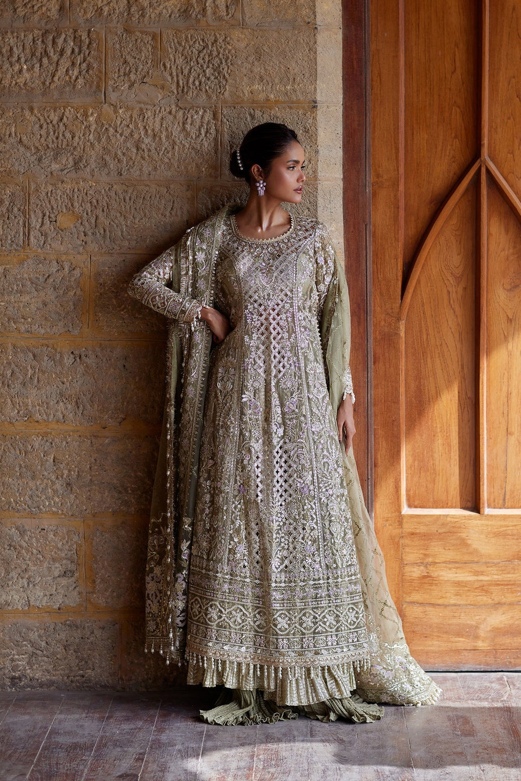 A woman models an intricately embroidered light beige and olive green traditional Anarkali gown with a matching dupatta, standing in a doorway next to a stone wall.