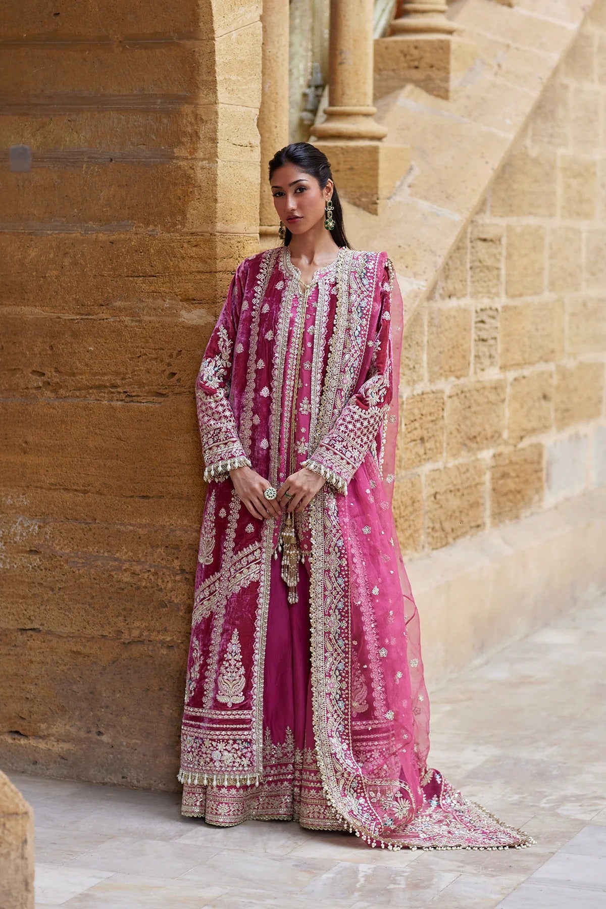A woman models a traditional Indian outfit consisting of a magenta pink kurta and matching pants with extensive gold embroidery, paired with a matching sheer dupatta, set against a warm stone archway background.