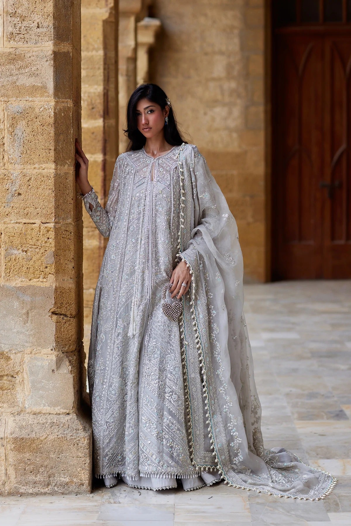 A woman wearing a long silver embroidered traditional outfit with a sheer matching dupatta, standing in a stone corridor with warm beige walls and a dark wooden door in the background.