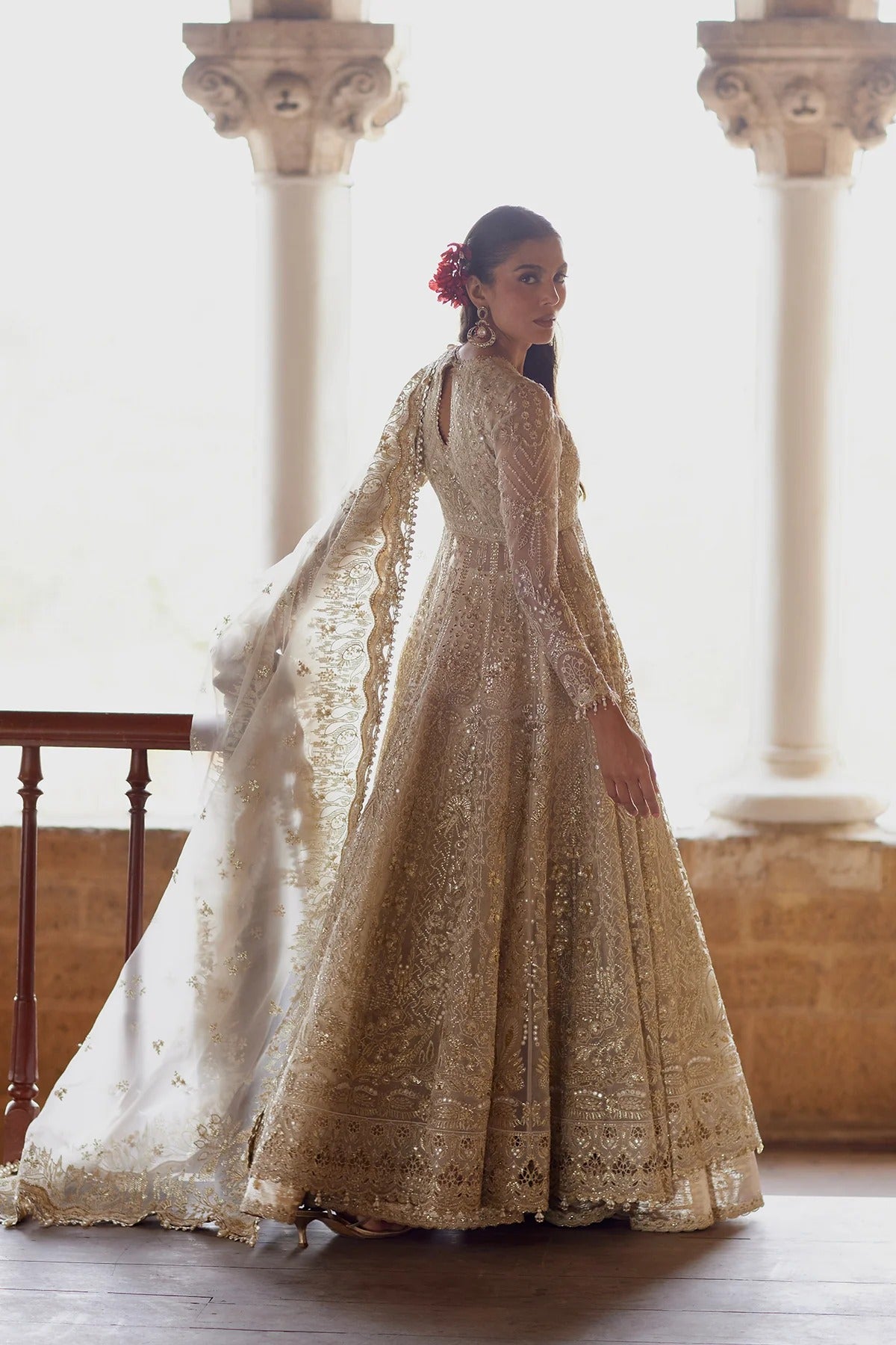 A woman wearing an ornate cream and gold embroidered traditional gown with a long draped dupatta, standing in a sunlit interior with stone columns.