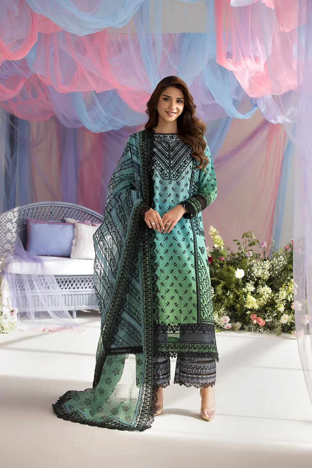 Woman in a green and black traditional outfit standing in a decorated room with colorful drapes and flowers.