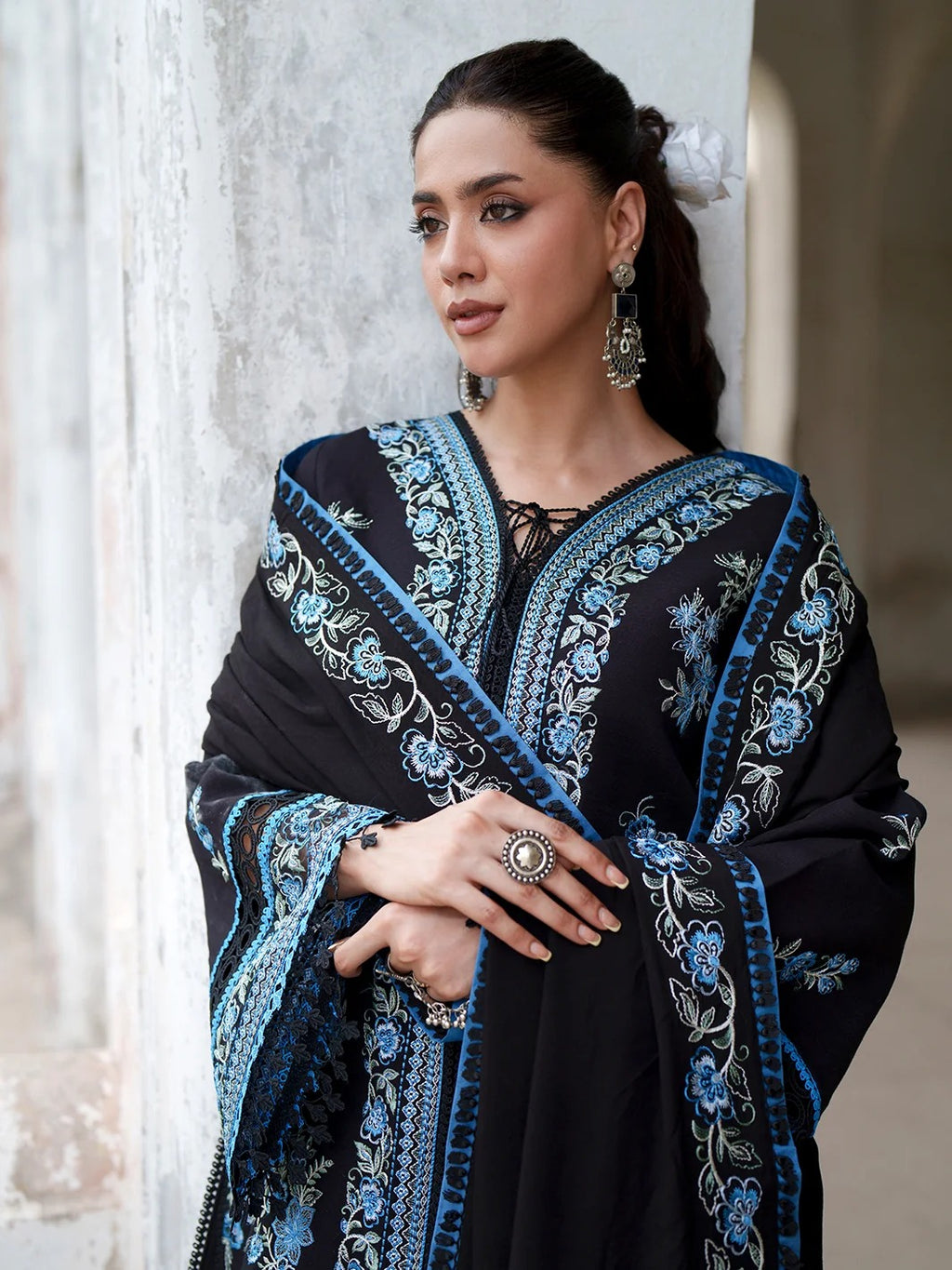 A woman wearing a black embroidered shawl or dupatta with intricate blue and silver floral patterns, posed against a textured light gray stone wall.