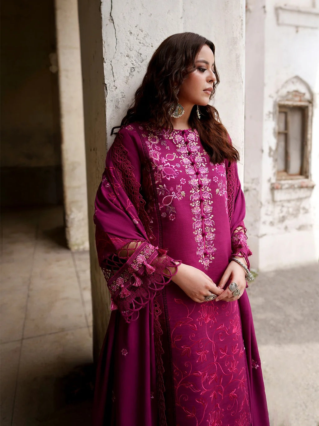 A woman wearing a deep magenta traditional embroidered dress with lace sleeves, standing in a rustic courtyard setting.