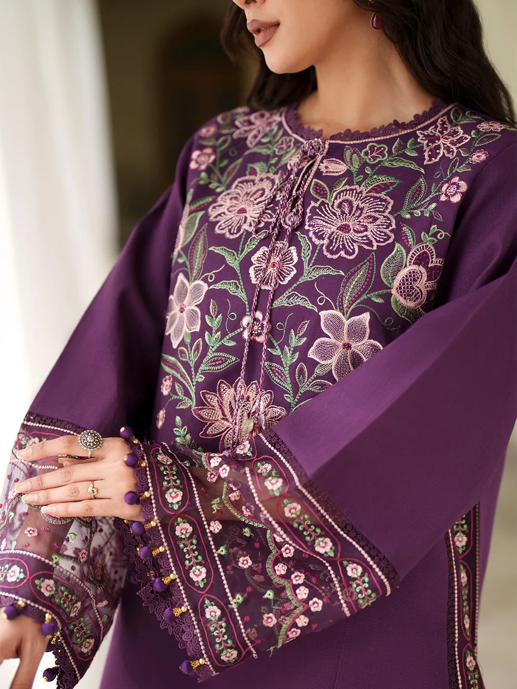 A close-up of a woman wearing a purple embroidered traditional outfit with floral embroidery in pink, green, and white, featuring sheer bell sleeves with tassel trim.
