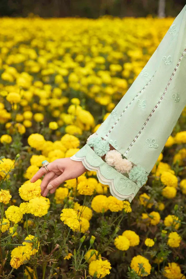 A close-up of a person's hand and arm wearing a light mint green embroidered sleeve, reaching toward bright yellow flowers in a field.