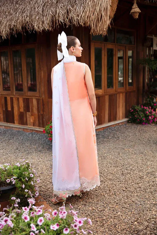 A woman stands outdoors in front of a wooden building with a thatched roof, wearing a peach and white traditional dress with a white headpiece. Pink flowers and gravel are visible in the foreground and background.