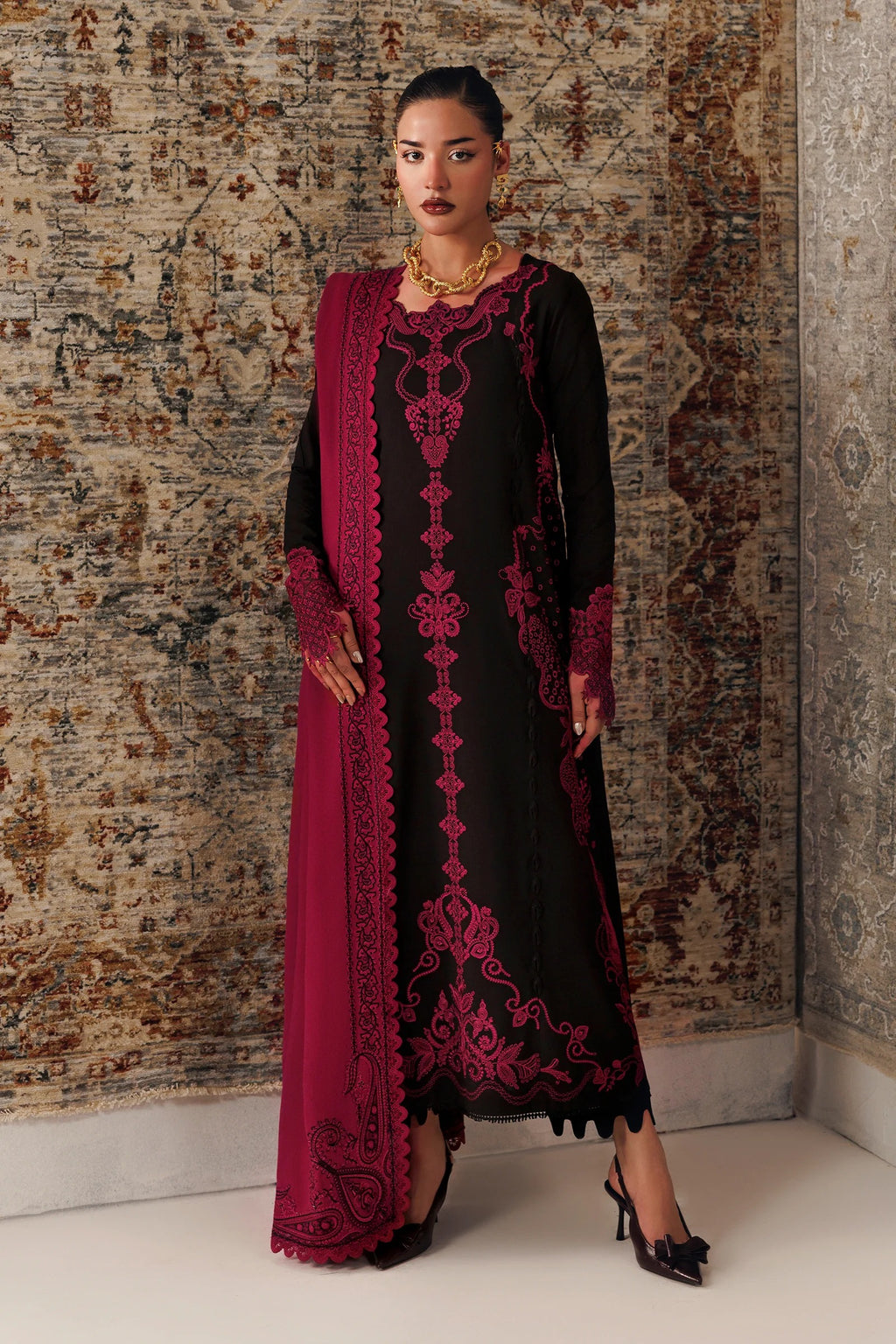 A woman models a black long kurta with deep magenta embroidery and a matching magenta dupatta, standing against a patterned brown and beige rug.