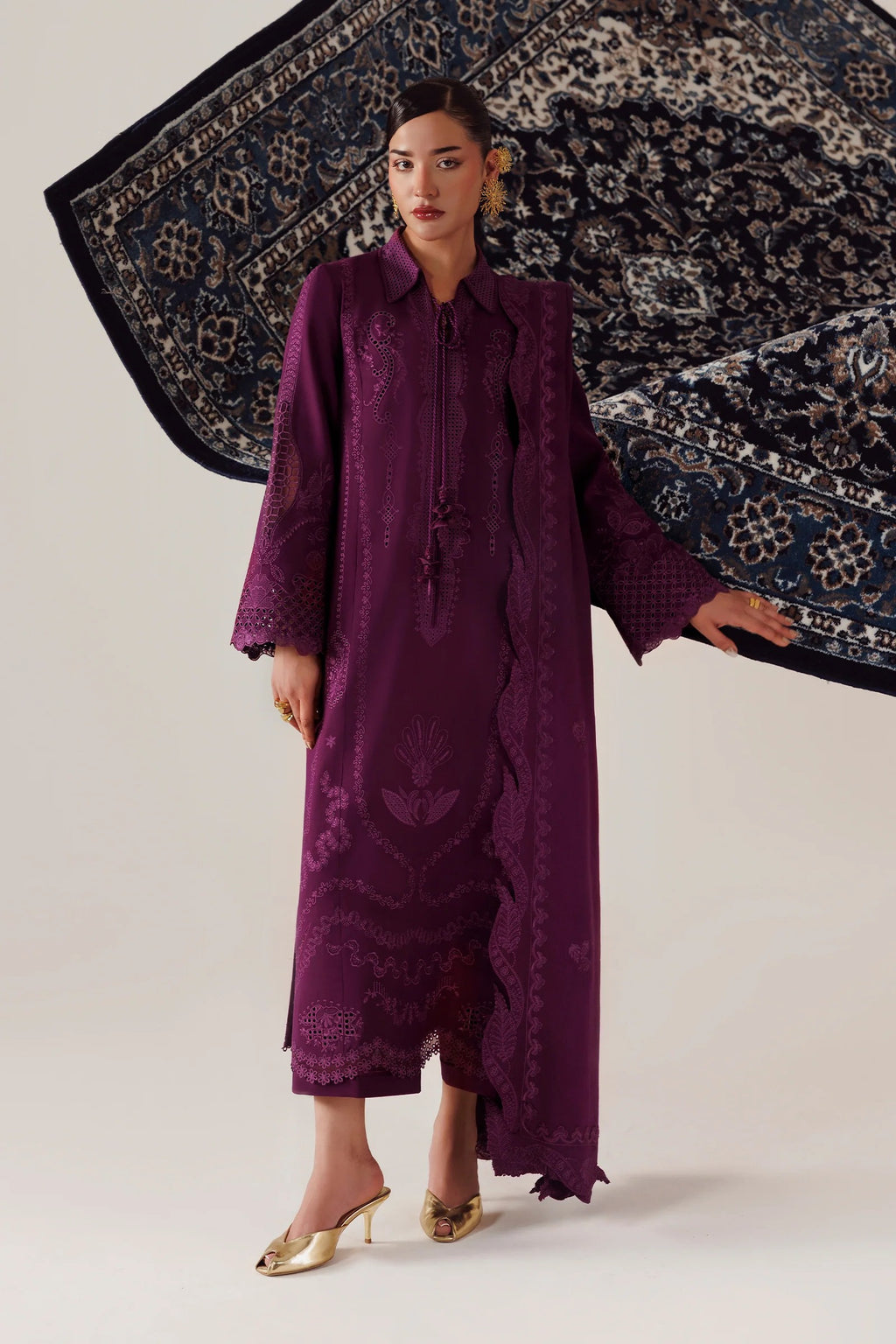 A model wearing a long, deep purple embroidered dress with gold heels, standing against a light beige studio background while holding a large patterned navy blue and beige rug.