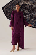 A model wearing a long, deep purple embroidered dress with gold heels, standing against a light beige studio background while holding a large patterned navy blue and beige rug.