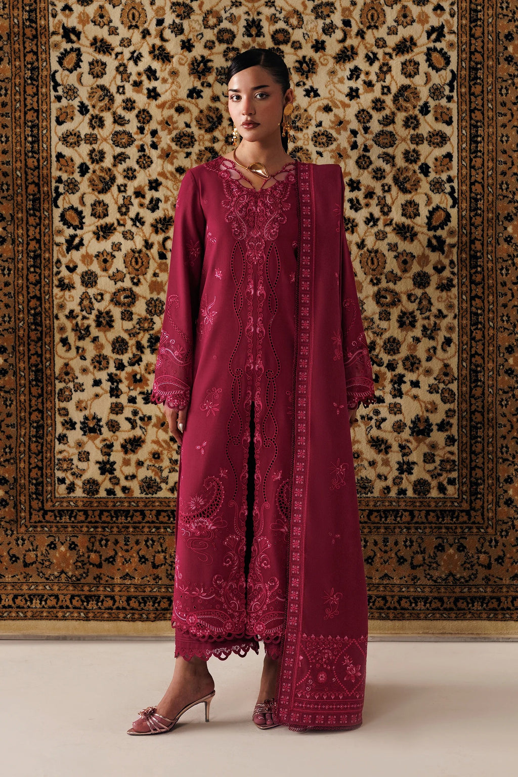 A woman models a deep magenta embroidered traditional outfit with a matching dupatta, standing in front of a large patterned oriental rug.