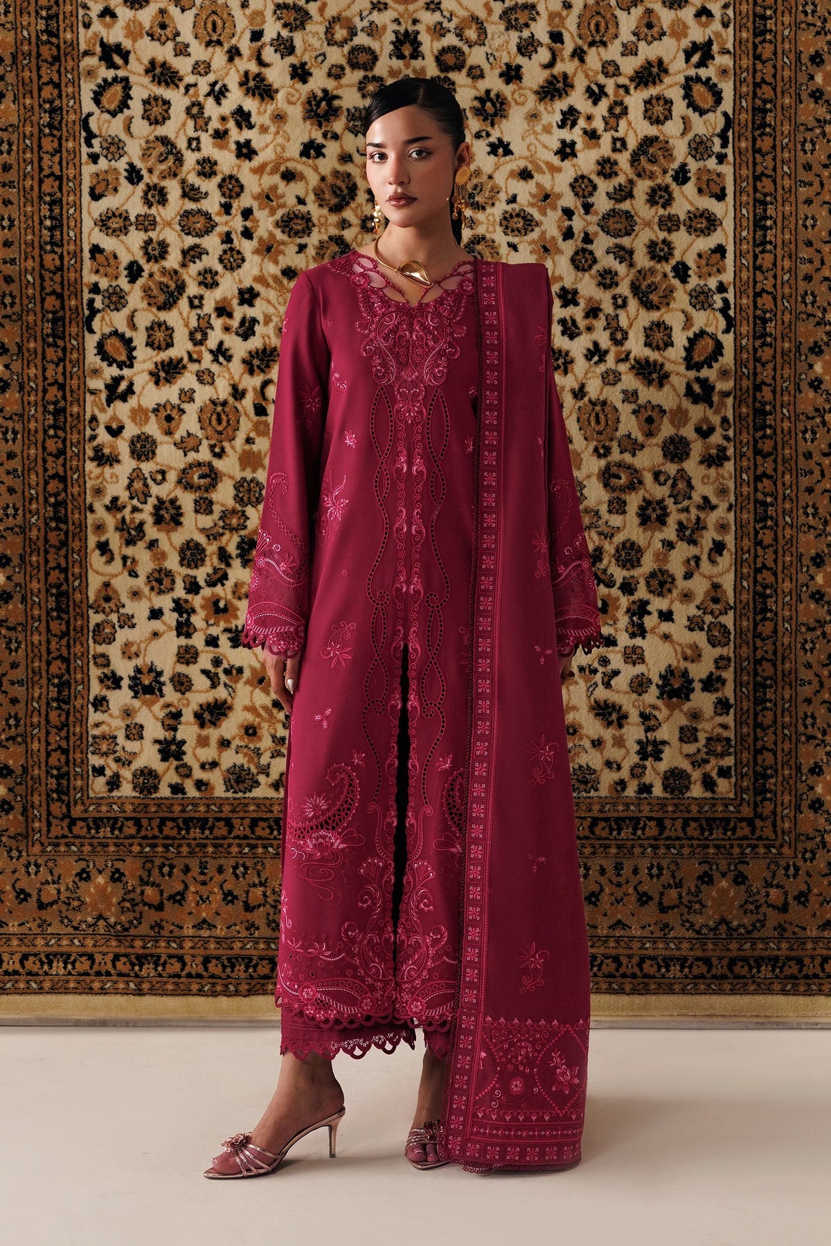 A woman models a deep magenta embroidered traditional outfit with a matching dupatta, standing in front of a large patterned oriental rug.