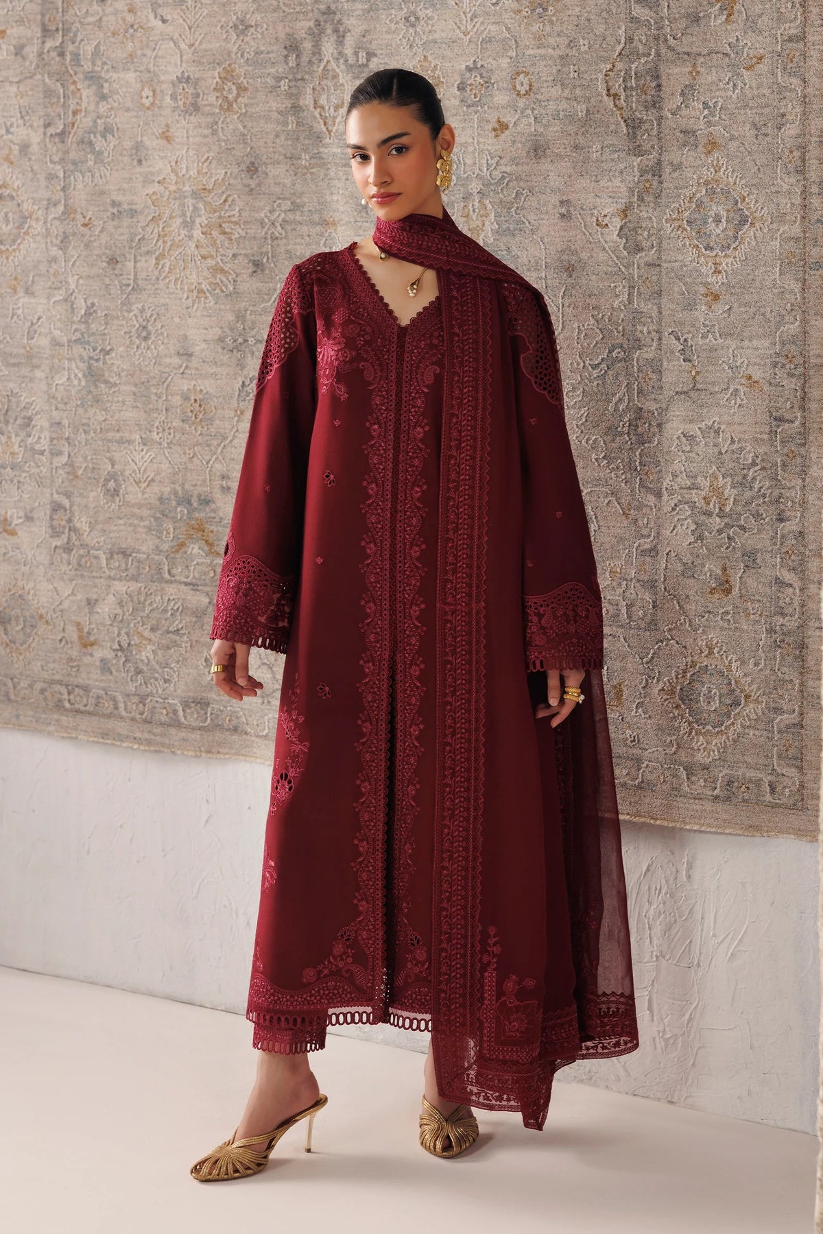 A woman models a long maroon embroidered traditional dress with a matching dupatta and gold heels, standing against a patterned beige wall.
