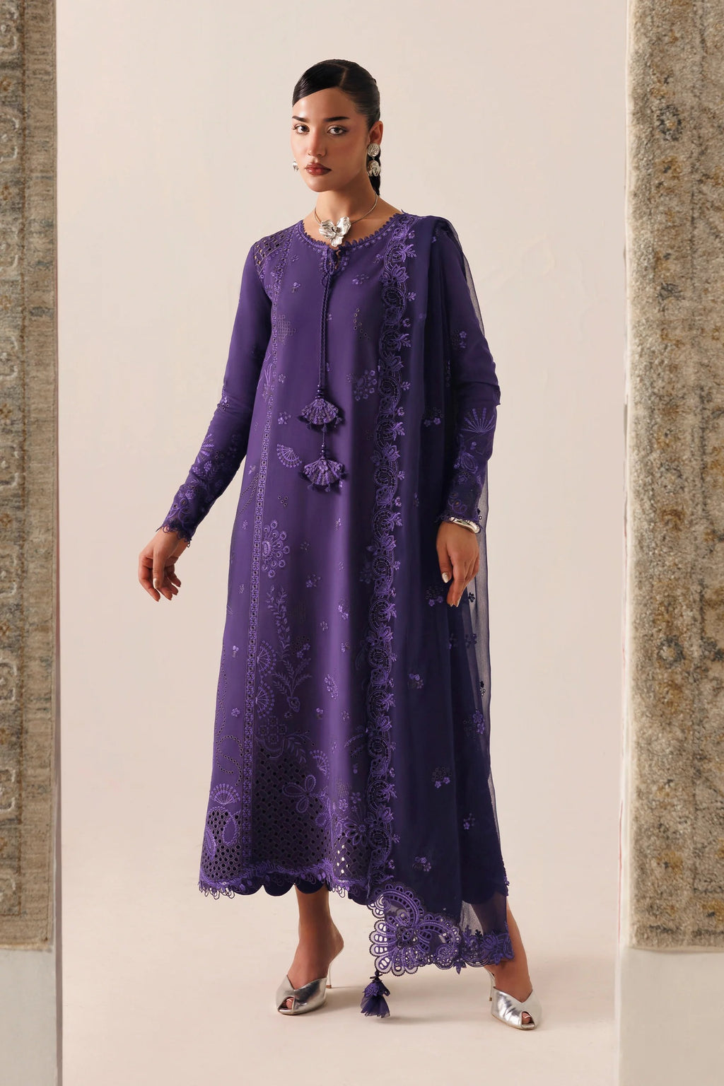 A woman models a long-sleeved purple embroidered kurta with a matching sheer dupatta, standing between two stone pillars against a light beige background.