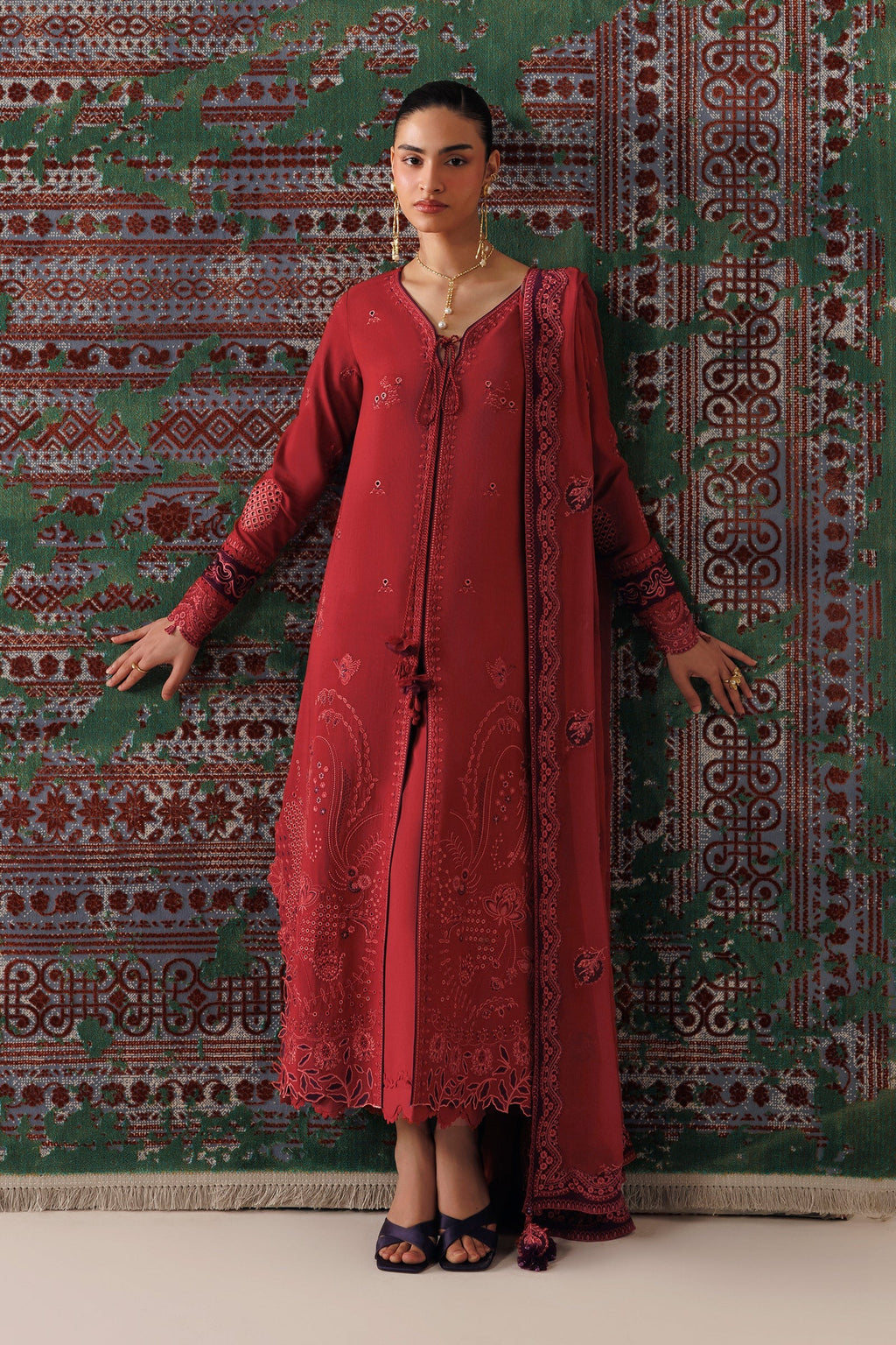 A woman models a traditional red embroidered kurta set with a matching dupatta, standing against a patterned green and red rug.