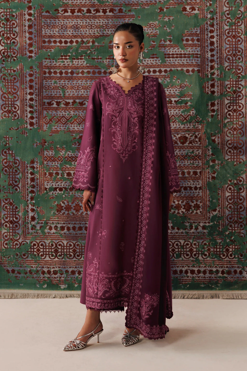 A woman models a long-sleeved, deep purple embroidered kurta with matching embellished border details, standing in front of a patterned green and red tapestry backdrop.