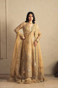 A woman models a traditional beige and gold embroidered Anarkali gown with a matching sheer dupatta, set against a neutral beige wall.
