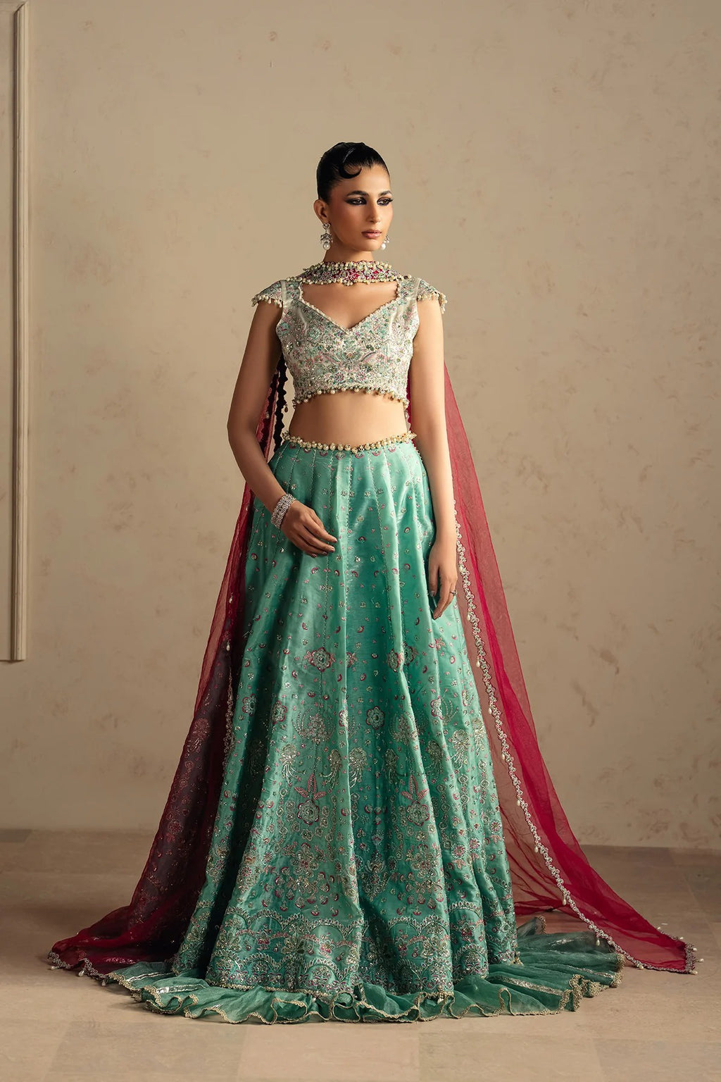 A woman models an ornate traditional Indian lehenga choli set. The outfit features a mint green embroidered skirt with floral patterns, a matching embroidered crop top, and a deep red dupatta draped over her shoulder and floor.
