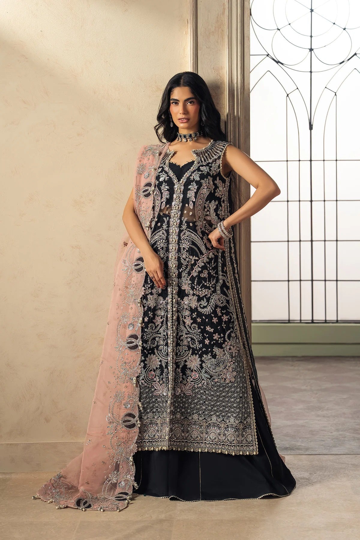 A woman models a black and silver heavily embroidered Anarkali gown with a sheer pink dupatta draped over one shoulder, set against a warm beige interior wall and window.
