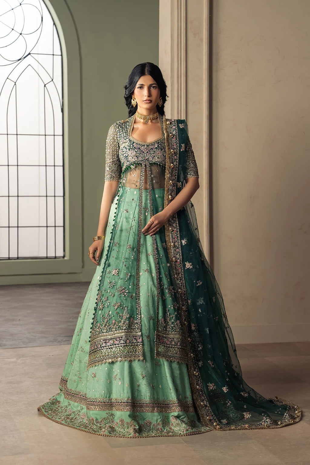 A woman models a traditional Indian lehenga choli set in a grand interior. The outfit is a mint green flared skirt (lehenga) with intricate gold embroidery, paired with a matching embroidered blouse and a dark teal dupatta.