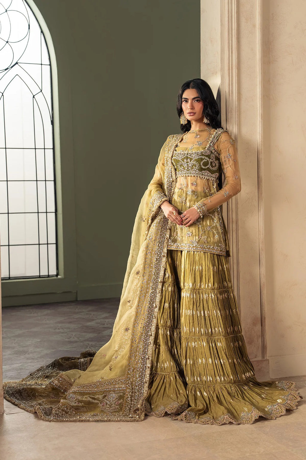 A woman models an ornate traditional Indian outfit consisting of a heavily embroidered olive green and gold lehenga skirt, a matching embellished blouse, and a long dupatta, posed in an elegant indoor setting with a large arched window.