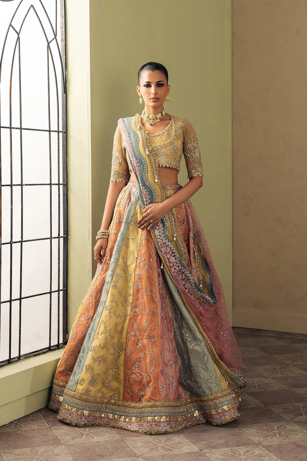 A woman models an ornate traditional Indian lehenga choli set in a styled indoor setting. The outfit features a heavily embroidered gold blouse with sheer sleeves, a voluminous skirt in a blend of peach, orange, and dusty pink with intricate gold and silver embroidery, and a matching dupatta draped over one shoulder.