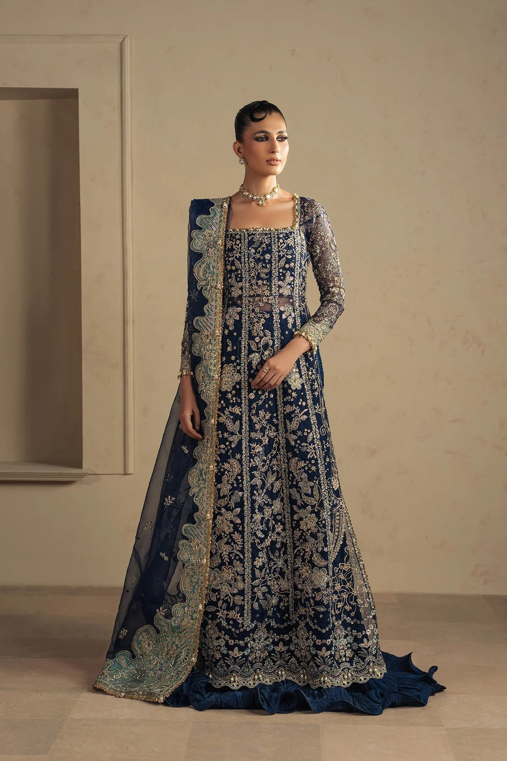 A woman models a traditional navy blue and gold heavily embroidered Anarkali gown with a matching sheer dupatta in a minimalist beige interior setting.