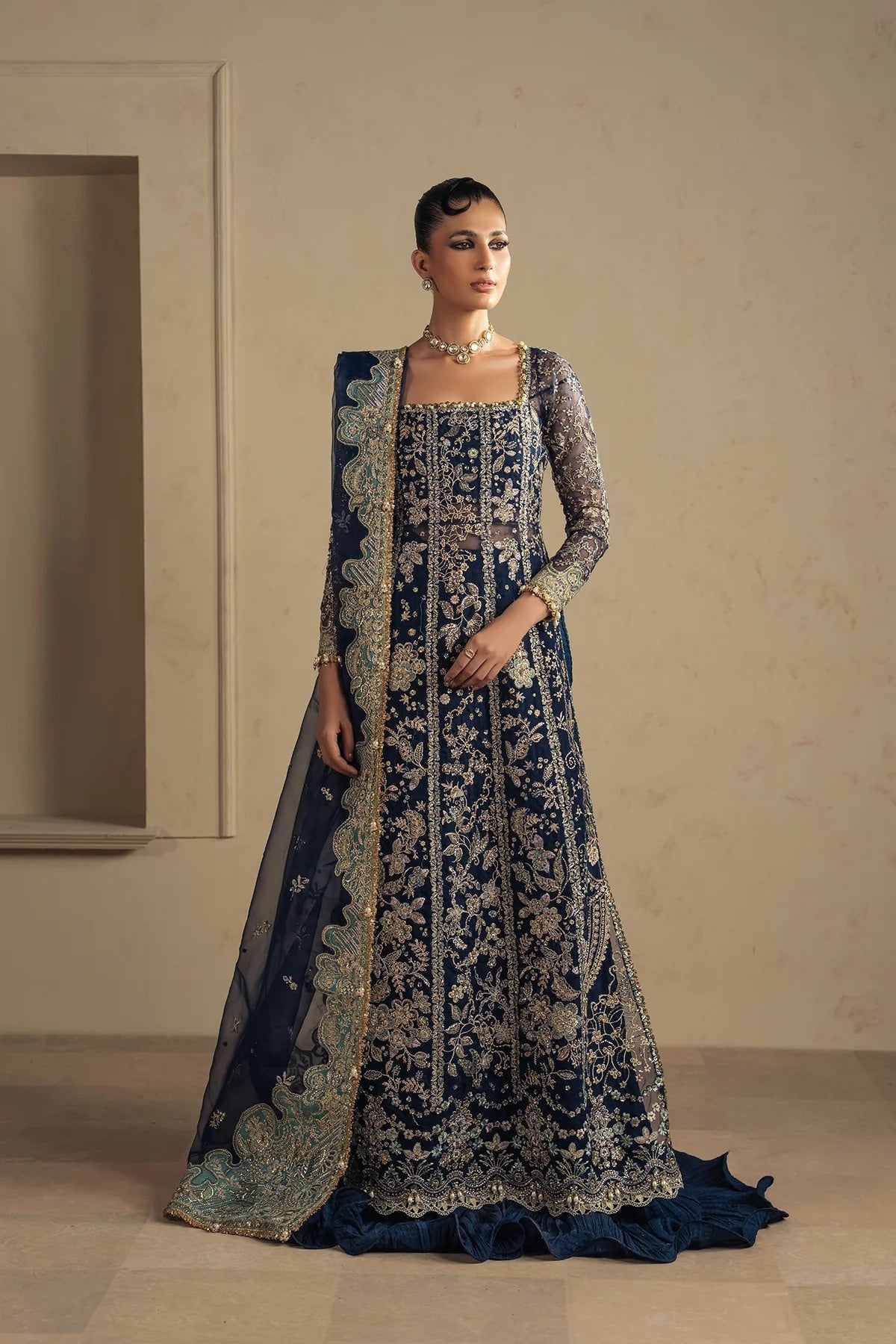A woman models a traditional navy blue and gold heavily embroidered Anarkali gown with a matching sheer dupatta in a minimalist beige interior setting.
