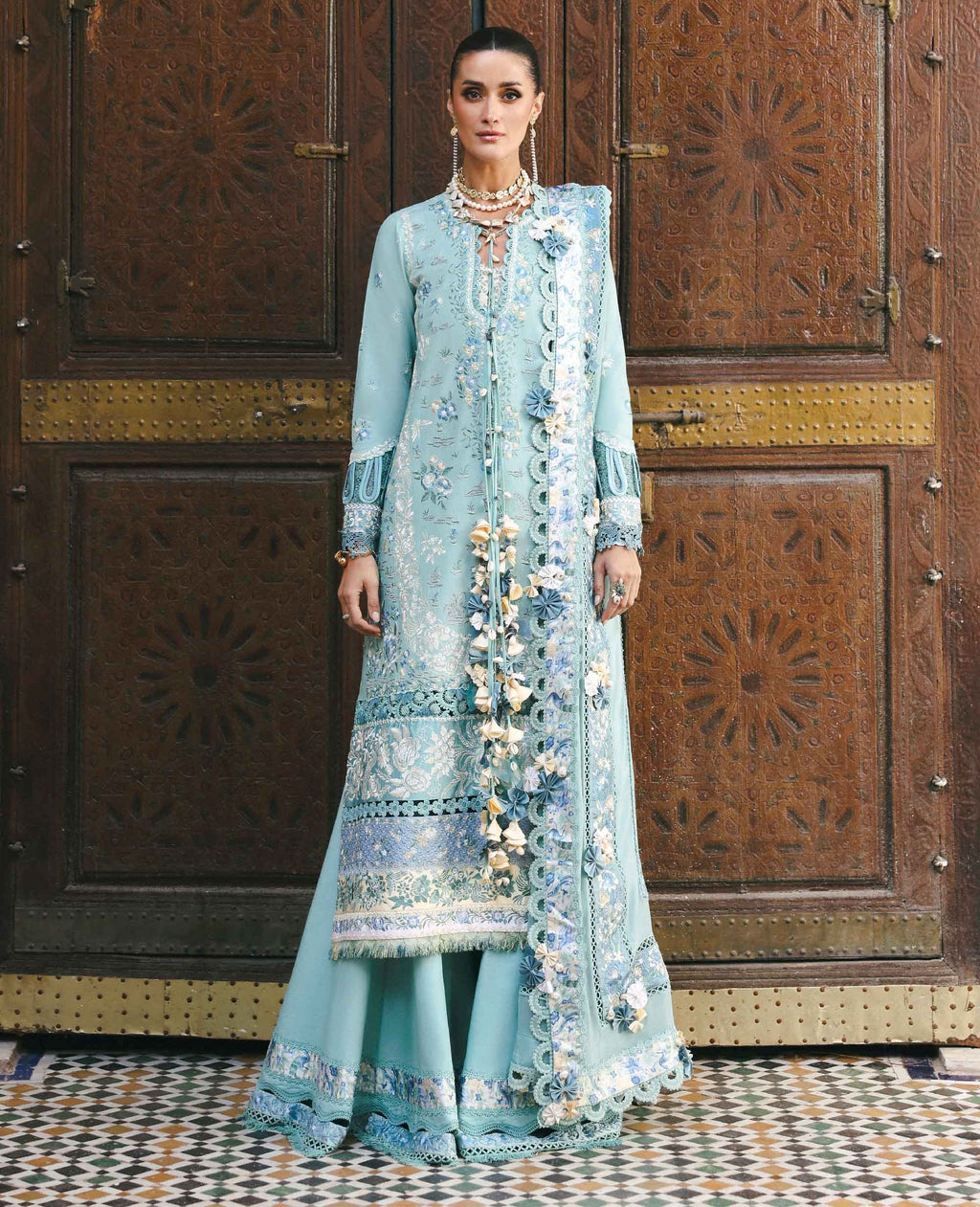 Woman in a light blue embroidered traditional outfit standing in front of an ornate wooden door.