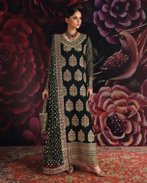 A woman models a traditional black embroidered kurta set with intricate gold and silver mirror-work patterns, set against a dark floral backdrop and a red patterned rug.