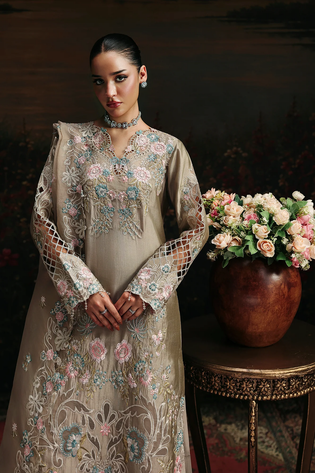 A woman models a beige embroidered traditional dress with floral patterns and sheer mesh sleeves, standing next to a wooden table holding a large vase of pink and white roses against a dark, painterly background.