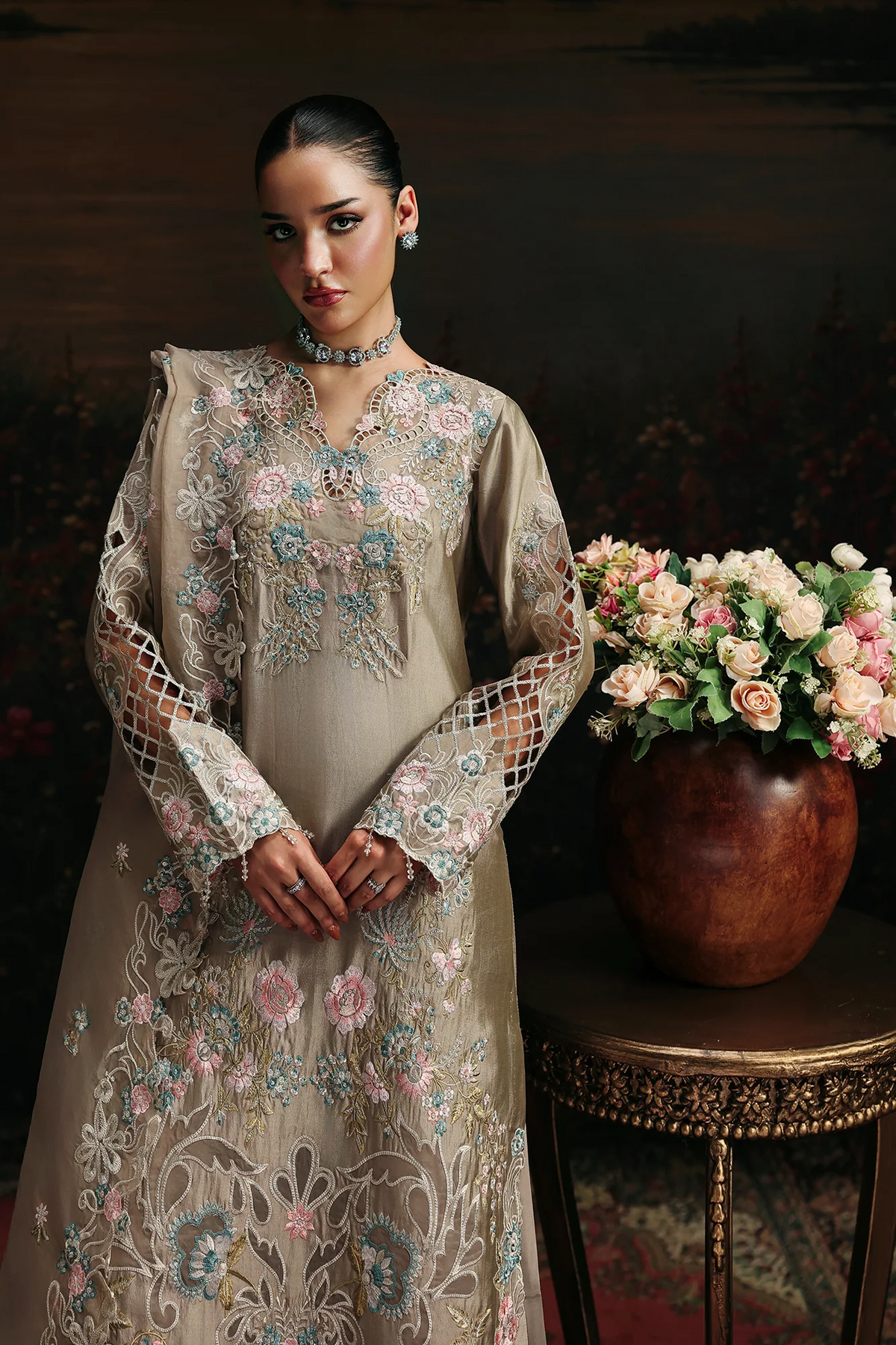 A woman models a beige embroidered traditional dress with floral patterns and sheer mesh sleeves, standing next to a wooden table holding a large vase of pink and white roses against a dark, painterly background.