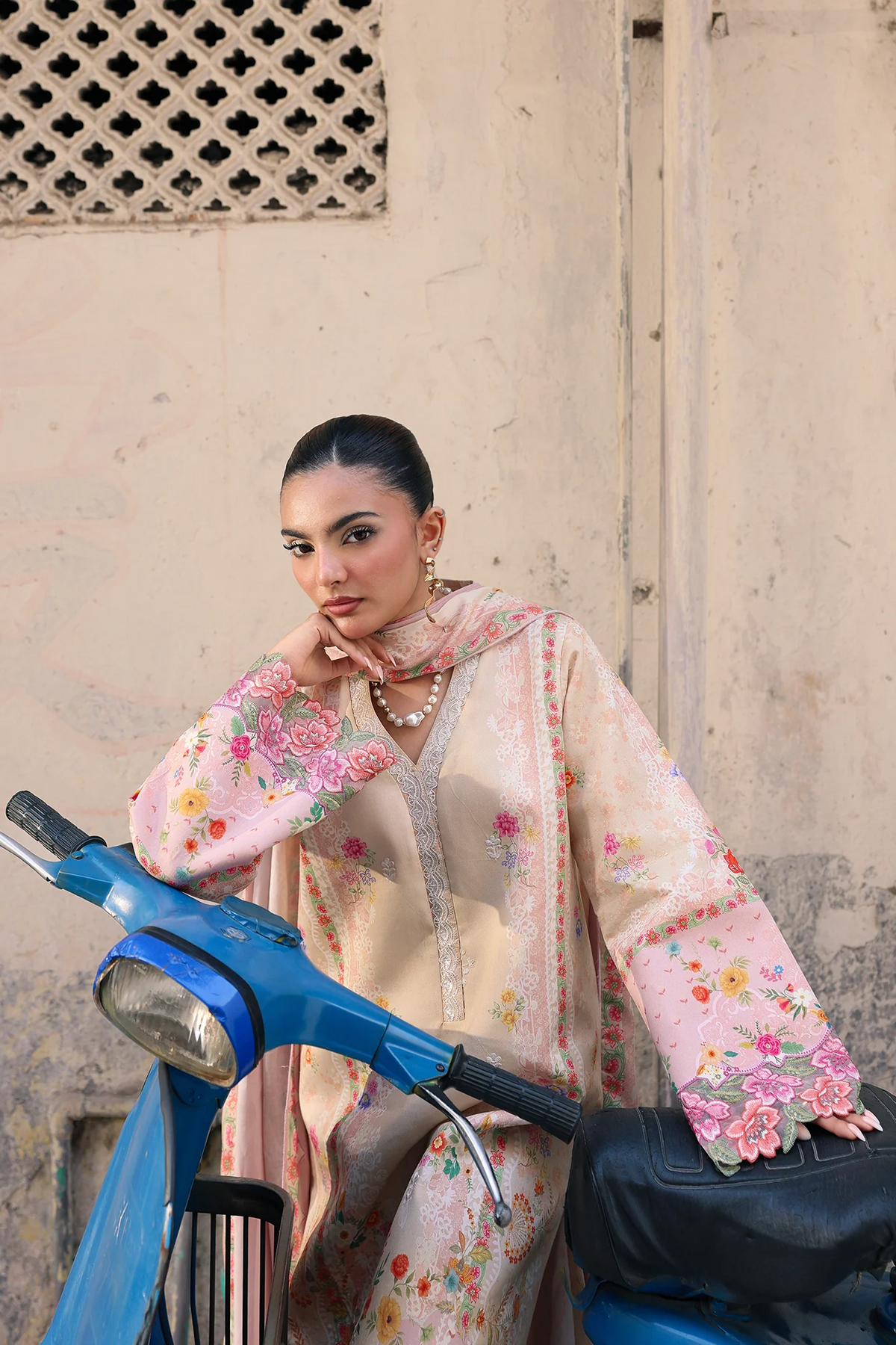 A woman wearing a light pink and beige floral embroidered abaya and matching scarf leans against a blue scooter in front of a textured beige wall.
