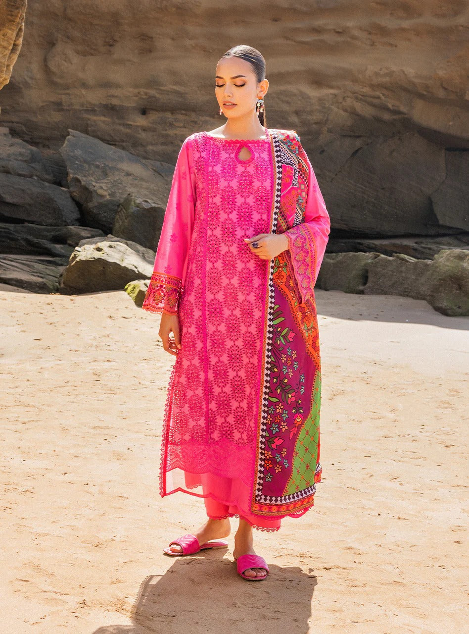 A woman stands outdoors on sandy ground in front of rock formations, wearing a bright pink traditional outfit with a matching printed dupatta. The outfit has ornate white and gold embroidery, and the dupatta features multicolor floral patterns with green, orange, and red sections.