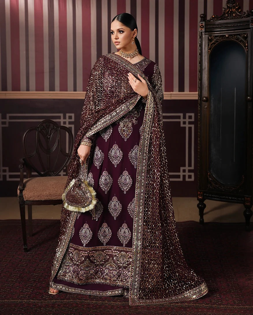 A woman models a heavily embellished traditional Indian outfit in a decorated room. The outfit is a deep maroon/burgundy lehenga with intricate silver and gold embroidery, a matching dupatta, and a small matching handbag.