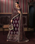 A woman models a heavily embellished traditional Indian outfit in a decorated room. The outfit is a deep maroon/burgundy lehenga with intricate silver and gold embroidery, a matching dupatta, and a small matching handbag.
