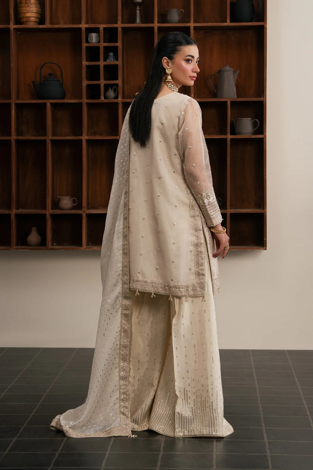 A woman stands with her back to the camera wearing a light beige traditional outfit with subtle embroidery and a matching sheer dupatta, posed in front of a dark wooden shelving unit.