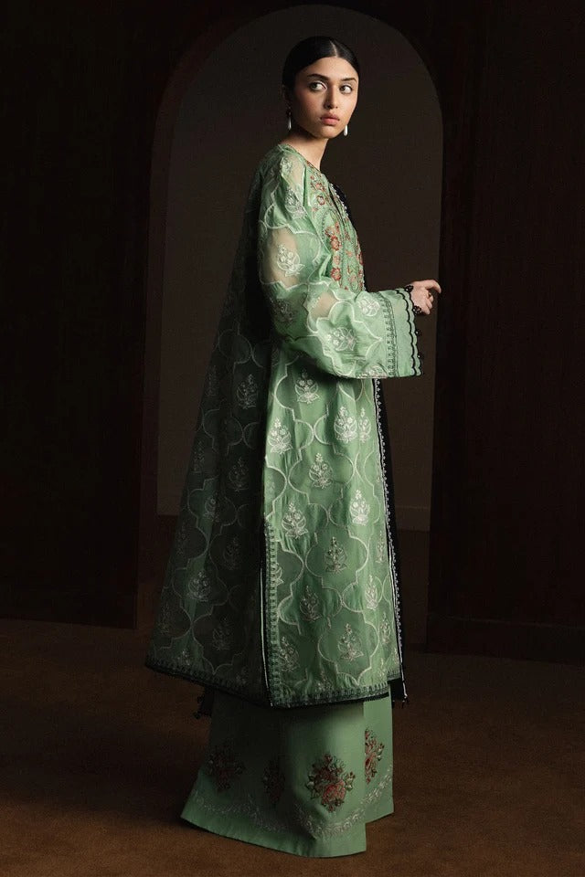 A woman models a traditional long green embroidered coat over a matching skirt in a dimly lit, arched doorway setting.