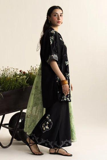 Woman in black and green traditional outfit standing next to a wheelbarrow with plants.
