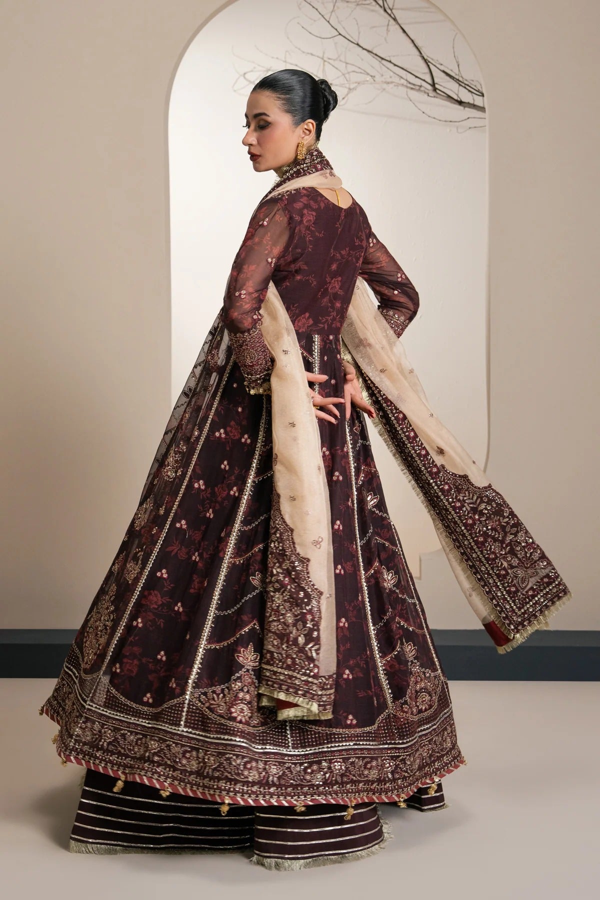 A woman modeling a traditional dark maroon embroidered Anarkali gown with a beige dupatta, shown from the back in a studio setting with a beige wall and arched backdrop.