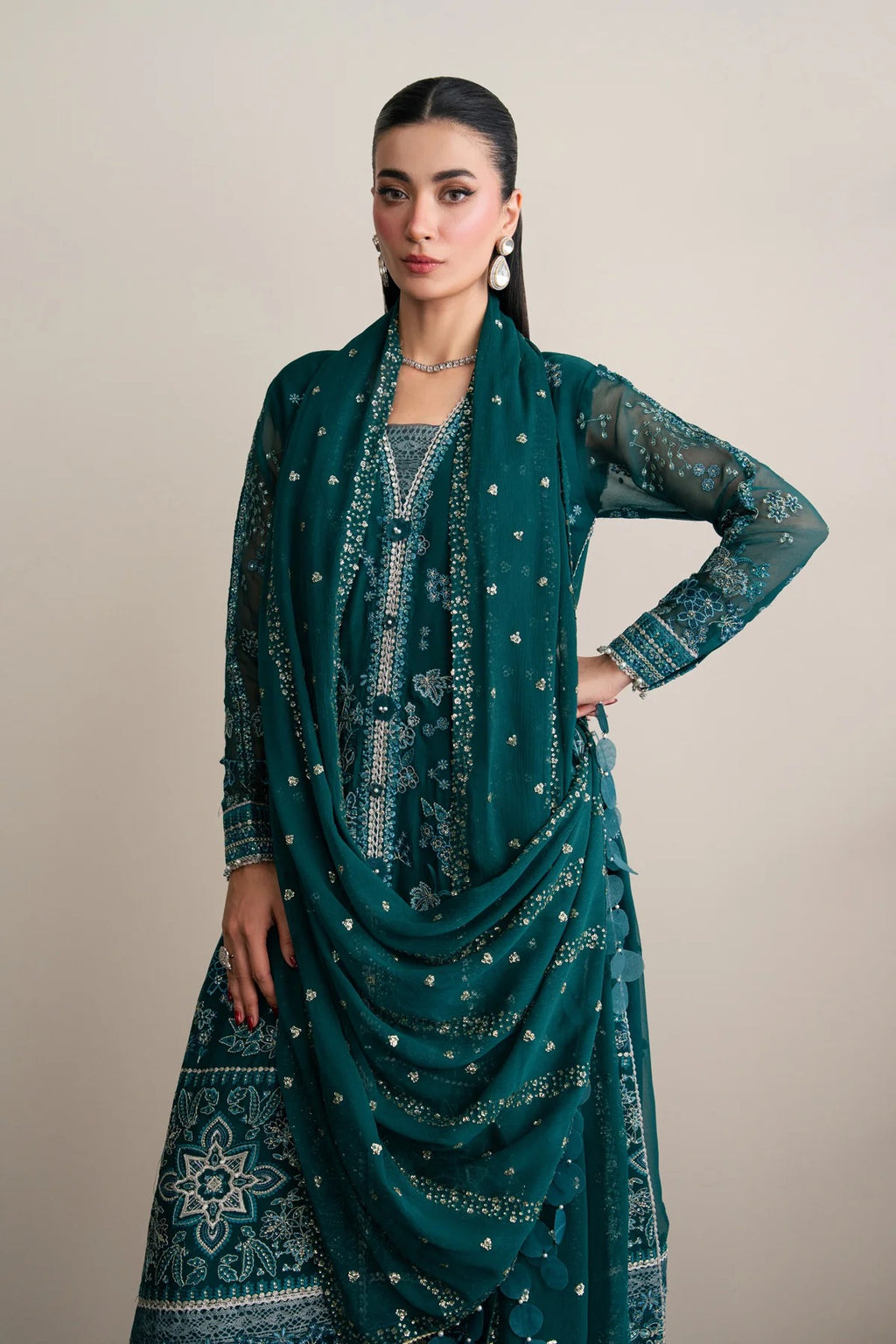 A woman models a dark teal green traditional South Asian outfit with intricate silver embroidery and a matching dupatta, set against a plain beige studio background.