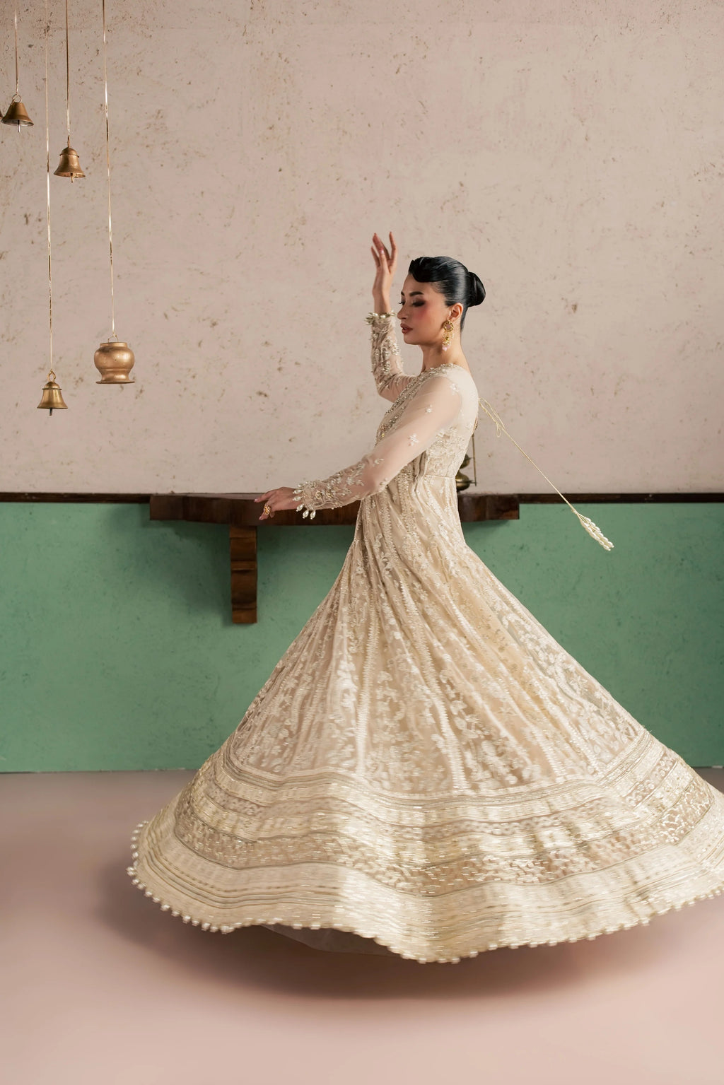 A woman wearing a cream-colored, heavily embroidered traditional anarkali-style dress twirls in a room with a textured beige and green wall.