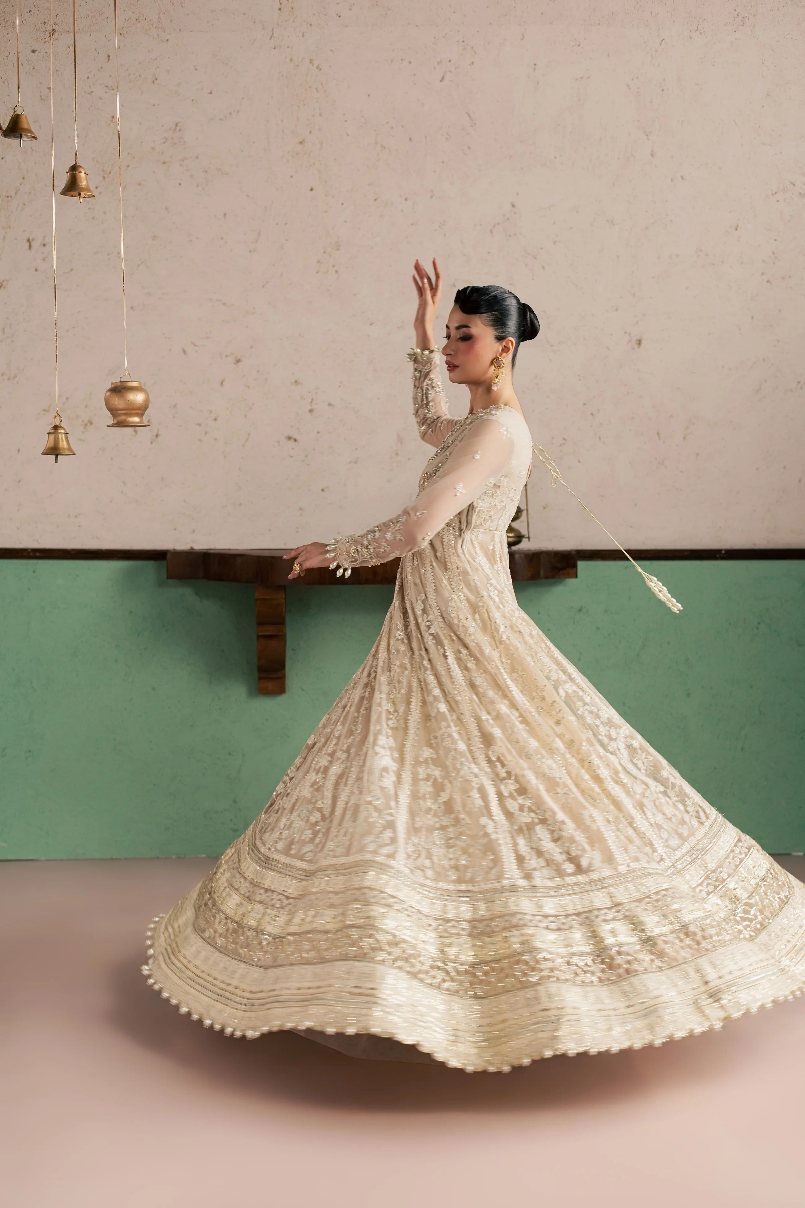 A woman wearing a cream-colored, heavily embroidered traditional anarkali-style dress twirls in a room with a textured beige and green wall.