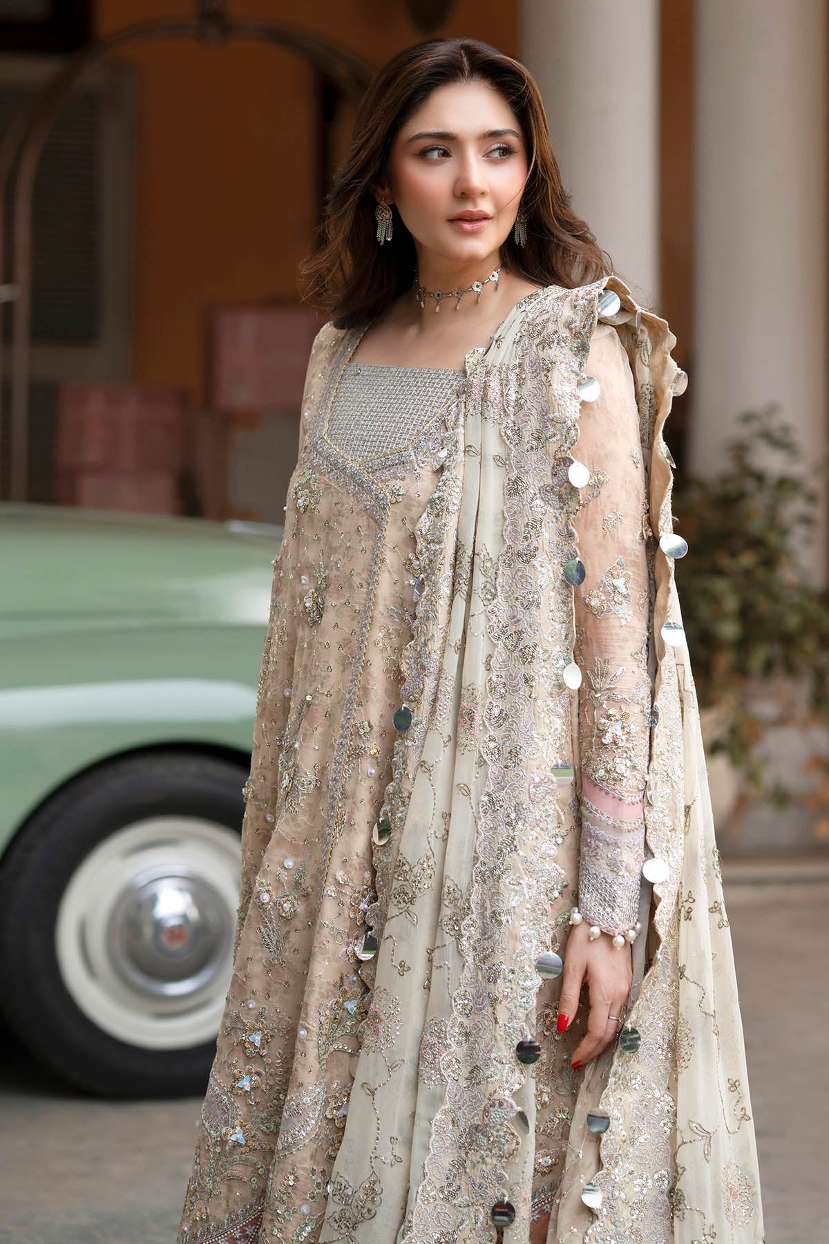 A woman wearing a traditional beige embroidered anarkali suit with a matching draped jacket and embellished dupatta, standing outdoors in front of a vintage green car.