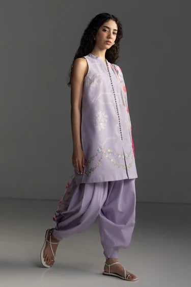 A woman modeling a light purple/lavender traditional outfit with matching loose pants and sandals, photographed against a plain gray studio background.