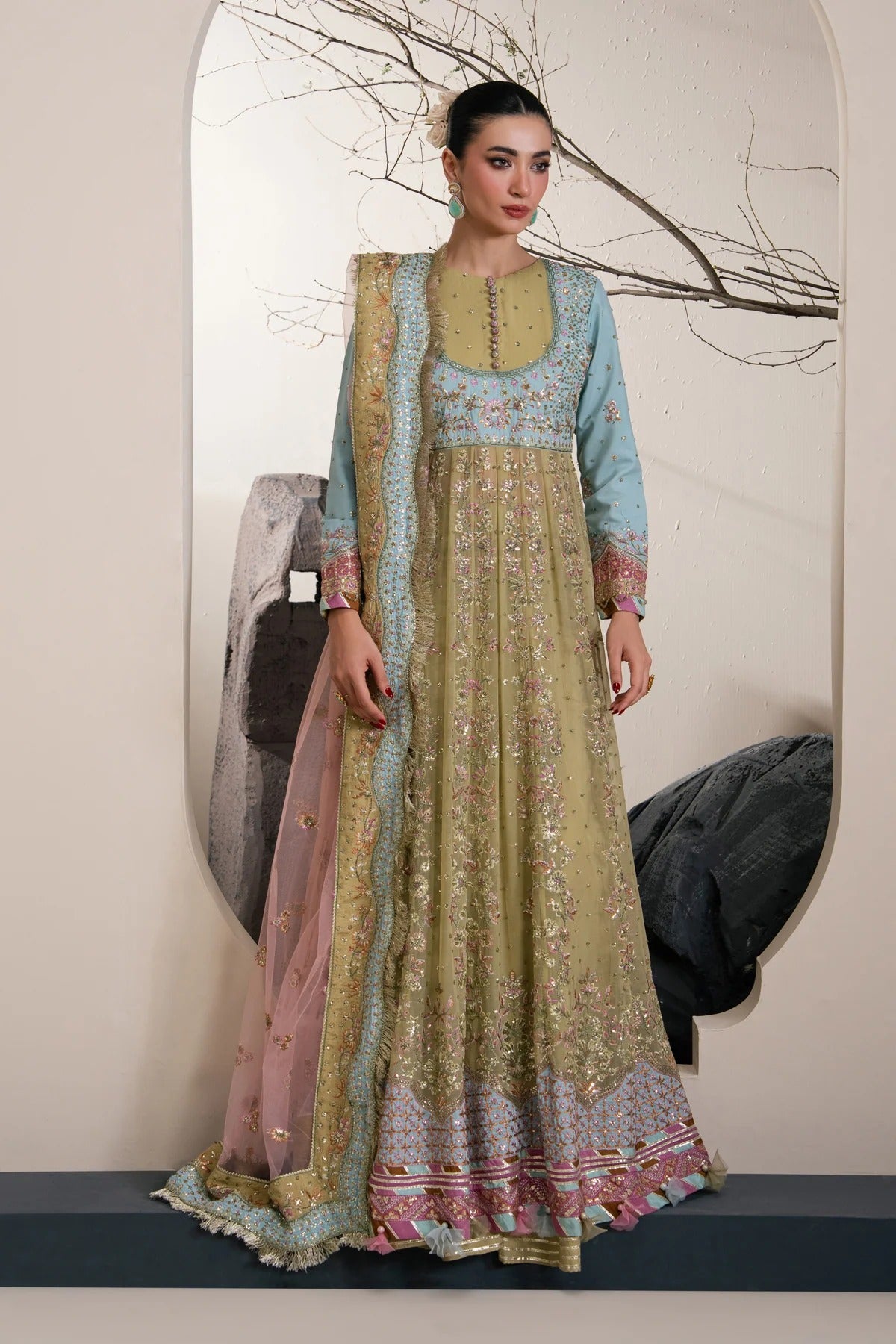 A woman models a traditional Indian salwar kameez outfit with a light blue kurta, a beige embroidered kameez, and a pink dupatta with gold detailing, set against an indoor backdrop with a large arched mirror.