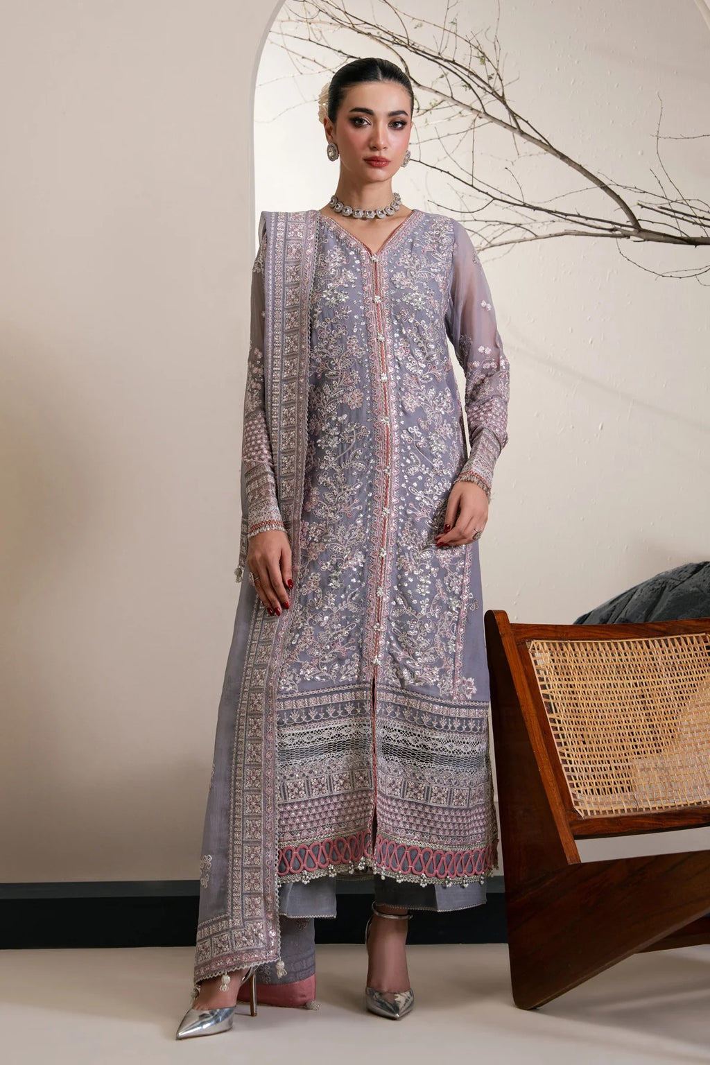 A woman models a traditional three-piece salwar kameez suit in lavender with intricate silver and pink floral embroidery, standing in a styled indoor setting.