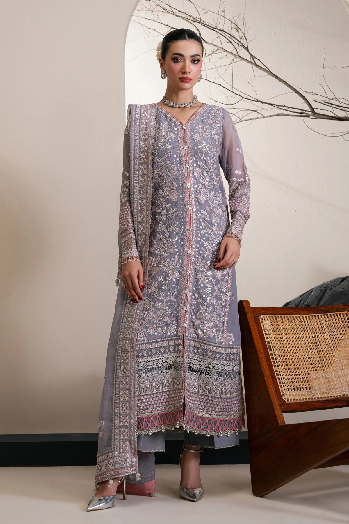 A woman models a traditional three-piece salwar kameez suit in lavender with intricate silver and pink floral embroidery, standing in a styled indoor setting.