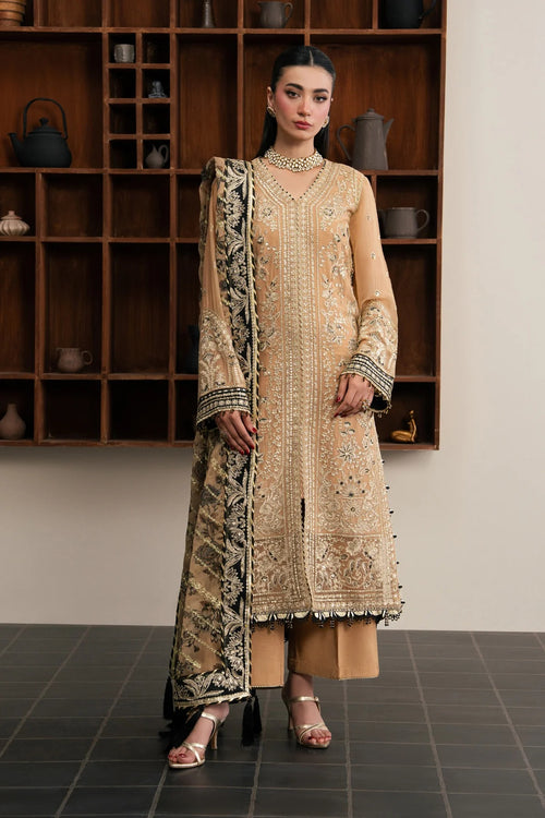 A woman models a traditional beige and gold embroidered salwar kameez suit with a matching printed dupatta, standing in front of a dark wooden shelving unit.