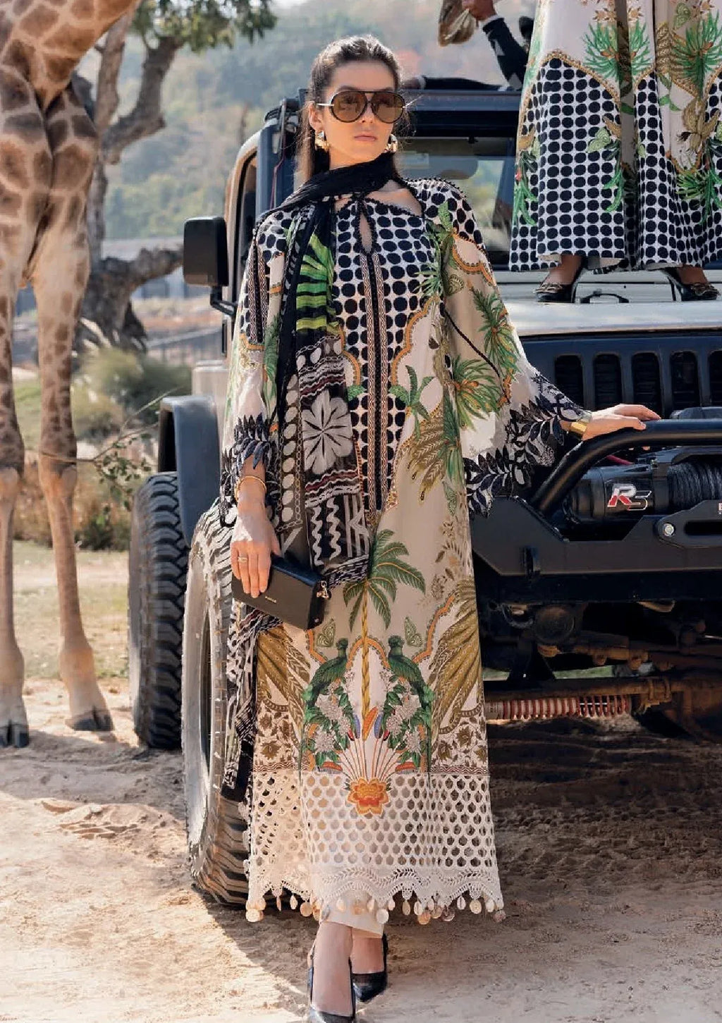 A woman wearing a long beige and black patterned kaftan dress with green botanical prints stands outdoors next to a black off-road vehicle, with a giraffe visible on the left.